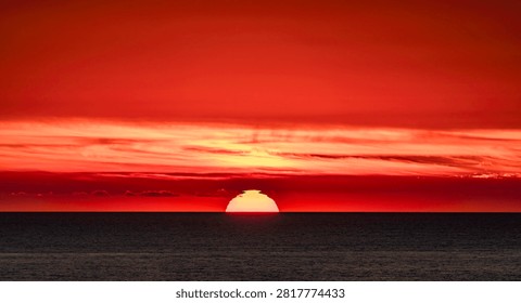 The sun dips below the ocean's edge, painting the sky with fiery hues and wispy clouds