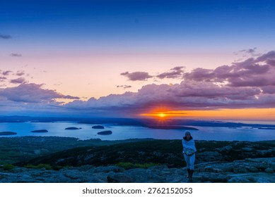 Sun dips low over distant islands. Sky glows with purple, orange, and soft blue hues. Lone figure stands on rocky peak, gazing at horizon. Silhouetted against warm glow, they appear serene