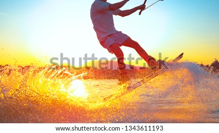 SUN FLARE, CLOSE UP: Cool surfer dude does 180 ollie while wakeboarding on a sunny summer evening. Cinematic shot of a cheerful man wakeboarding in the cool cable wake park. Awesome summer water sport
