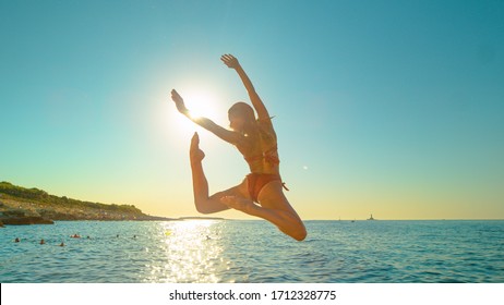 SUN FLARE: Energetic Girl Jumps Off A Cliff And Kicks Legs Back While Diving Into The Ocean On A Sunny Summer Evening. Carefree Young Woman Does An Attractive Dive Into The Refreshing Sea At Sunrise.