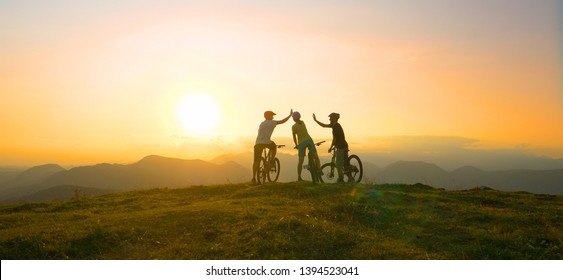 SUN FLARE: Mountain Biking Friends High Five After Reaching The Scenic Summit At Breathtaking Golden Sunrise. Cross Country Cyclists Celebrate A Successful Mountain Biking Adventure On A Sunny Evening