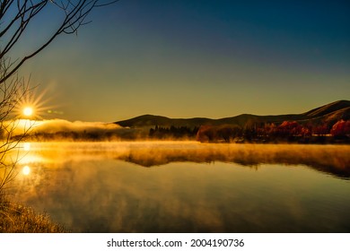 Sun flare and mountain reflections hovering above the water surface at the deserted lake at sunrise in the mist and low cloud