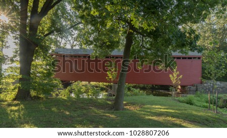 the sun forms a starbust throuhg the teee leaves at the Loyâ??s Station covered bridge near Thurmont MD. The red wooden bridge spans a stream during the summer green foliage.