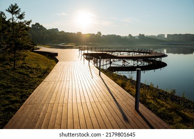 Sun glare at dawn of the day illuminates the embankment of the city park, the walking area, the wooden pier, the path of boards, the morning light, the pond embankment. High quality photo