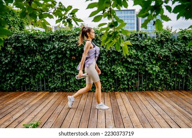 Sun glare effect. Active young female walking on wooden path outdoor at modern rooftop of building with green plants. Sport, healthy lifestyle concept.