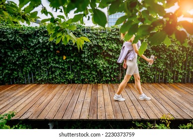 Sun glare effect. Active young female walking on wooden path outdoor at modern rooftop of building with green plants. Sport, healthy lifestyle concept. - Powered by Shutterstock