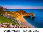 The sun goes down on the Jurassic coast and Durdle Door in Dorset with Seak Pink Trift growing on the top of the cliff in the foreground