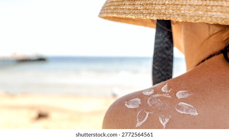 Sun hat shields shoulder from beach sun rays. White sunscreen forms flower pattern on skin. Sandy shore meets calm sea in soft focus. Person relaxes, enjoying summer holiday vibe