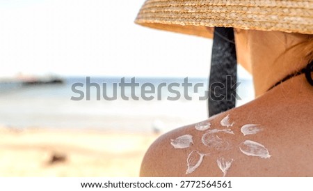 Sun hat shields shoulder from beach sun rays. White sunscreen forms flower pattern on skin. Sandy shore meets calm sea in soft focus. Person relaxes, enjoying summer holiday vibe