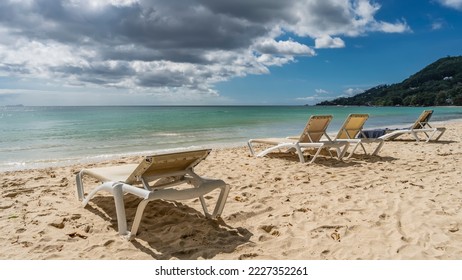 Sun loungers are on the sandy beach. Footprints all around.  The turquoise ocean is calm. A green hill against a background of blue sky and clouds. Seychelles. Mahe. Beau Vallon - Shutterstock ID 2227352261