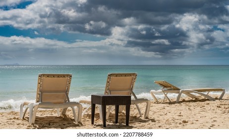 Sun loungers and a wicker table stand on the sandy beach. The turquoise ocean is calm. Foam of waves on the shore. Picturesque clouds in the blue sky. Seychelles. Mahe. Beau Vallon - Shutterstock ID 2207788459