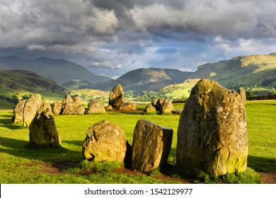 Sun on Castlerigg Stone Circle under dark clouds on summer solstice eve with Naddle village in Cumbrian Mountains Lake District Keswick England