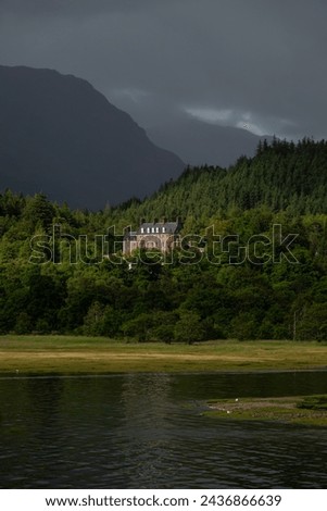 Sun peeks through dark clouds and illuminates beautiful mansion among trees. Secluded grand manor, standing at the foot of rugged mountains in Scottish Highlands, surrounded by forest and river.