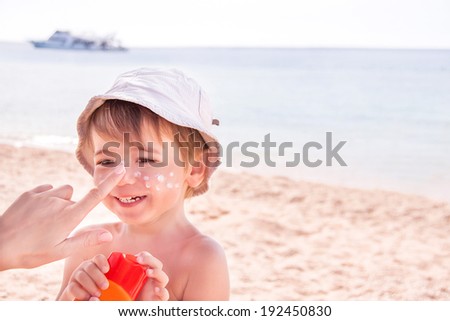 Sun protection. Hand of caucasian mother applying suncream  (suntan lotion) from a plastic container to her happy son before tanning during summer holiday on beach.  Copyspace, close up.