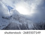 Sun rays break through thick clouds, illuminating the snow-covered mountain peak. Snowflakes swirl in the air, creating the atmosphere of a frosty winter day in the mountains