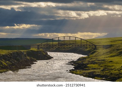 Sun rays breaking through clouds illuminate a metal bridge crossing a flowing river in the Icelandic countryside, creating a serene and dramatic scene.