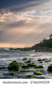 Sun rays breaking through clouds over a rocky coastline with moss-covered stones and gentle waves, creating a peaceful and dramatic seaside landscape.