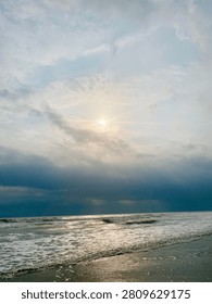 Sun rays breaking through clouds over ocean, clouds air scenery sun light blue sky. Beautiful landscape with golden sun lights penetrated through dark clouds. White water hitting the beach with small waves. Idea for natural, oceanic, and landscape themes.