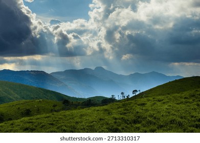 Sun rays breaking through dramatic clouds over lush green hills in Kerala, India. Scenic mountain landscape with rolling grasslands, natural light, and open space for background use.