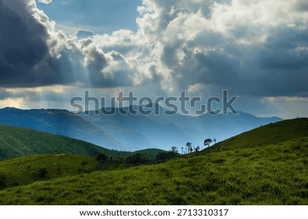 Sun rays breaking through dramatic clouds over lush green hills in Kerala, India. Scenic mountain landscape with rolling grasslands, natural light, and open space for background use.