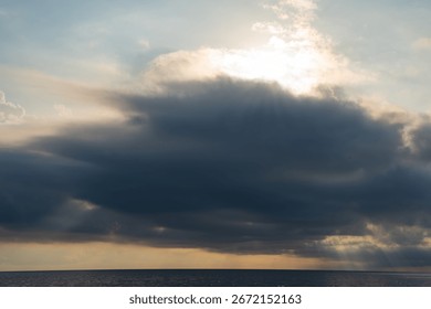 Sun rays breaking through large, dark cloud over calm ocean. Straight horizon separating calm sea from dramatic