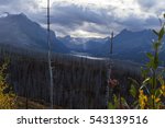 Sun rays breaking thru the clouds in a beautiful mountain landscape of the east side of Glacier National Park with St. Mary Lake and a section of dead timber from an old wildfire.