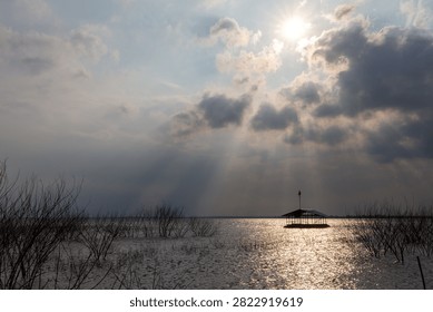 Sun rays piercing through clouds over a lake with a floating wooden raft house and submerged trees.
