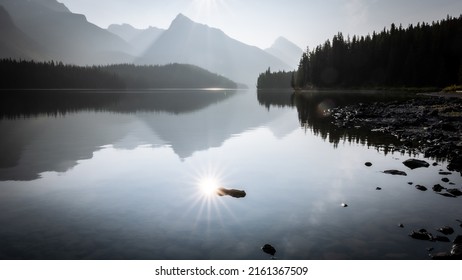 Sun reflecting in still alpine lake surrounded by mountains and forest, Jasper NP, Canada