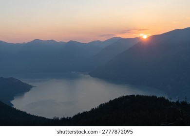 Sun rises behind mountains, casting light over a calm lake. Mist fills the valleys as trees stand against the early morning sky. This moment captures the beauty of nature.