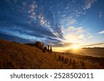 Sun rises over mountains, casting golden rays across landscape. Sky filled with dramatic clouds, creating stunning contrast with clear blue above. Warm light illuminates rolling hills and pine trees.