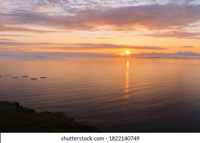 Sun Rising Behind Low Clouds At Sea In Scotland. Beautiful Orange Light Glow And Reflection In Water. Aquaculture Fishing Nets In The Sea