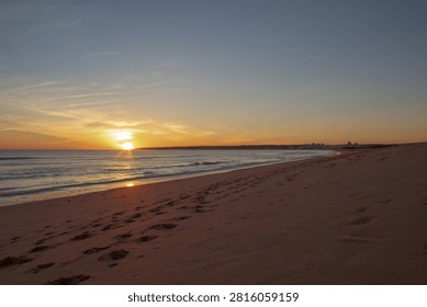The sun sets at Albufeira  over a beach, casting orange and yellow tones across the sky. Waves touch the shoreline, and footprints mark the sand. The scene is tranquil and inviting.