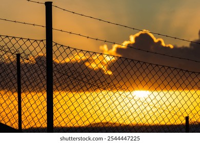 The sun sets behind a barbed wire fence, casting vibrant orange and yellow hues across the sky while clouds drift above, creating a striking evening atmosphere. - Powered by Shutterstock