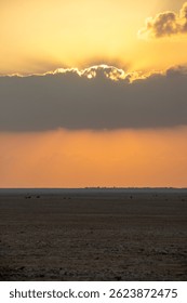 The sun sets behind clouds, casting golden rays over a vast, shadowy desert landscape at dusk.