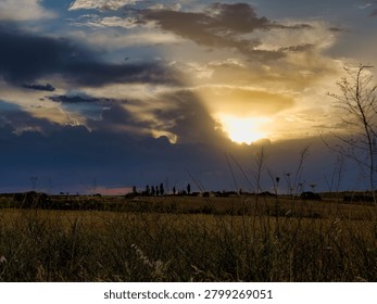 The sun sets behind clouds, casting light over a field. Silhouetted trees and hills appear against the colorful sky as shadows deepen in the grass.