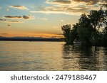 The sun sets behind the dam at Cherry Creek State Park in Colorado.  Blue, yellow, orange and red clouds show brilliance at the horizon. Dark blue water at the bottom of the image.