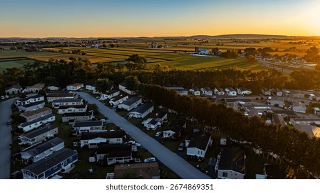 The sun sets behind distant hills, casting a warm glow over a peaceful mobile home residential neighborhood. Houses line the street, surrounded by lush green fields and farmland.