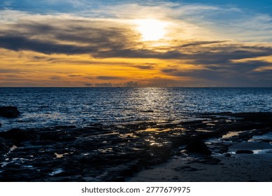 The sun sets behind a layer of clouds over the Indian Ocean, casting a golden glow on the water and a rocky beach in the foreground. Nosy Be, Madagascar