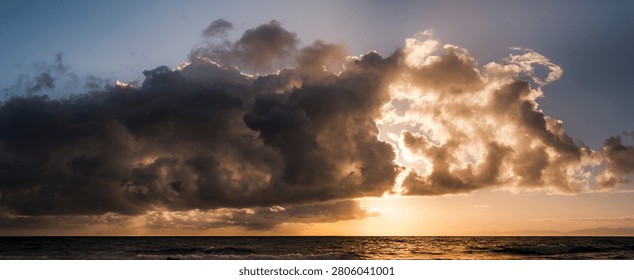 The sun sets behind a massive cloud formation over the sea, casting a warm glow on the water and sky