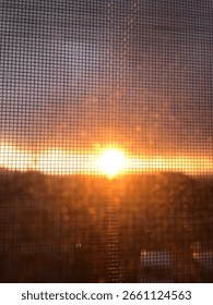 The sun sets behind a mesh screen, casting a warm golden glow across the sky and creating a soft, grid-like silhouette effect.