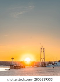 The sun sets behind a modern cable-stayed bridge, casting a warm glow over a sandy riverbank and calm water