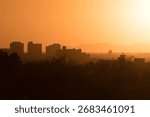 The sun sets behind mountains, casting a fiery orange glow across city skyline. Silhouetted buildings and faint outlines of trees merge with the hazy horizon, creating a dramatic desert sunset scene.