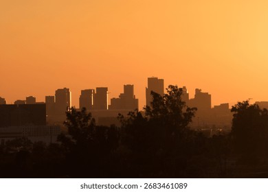 The sun sets behind mountains, casting a fiery orange glow across city skyline. Silhouetted buildings and faint outlines of trees merge with the hazy horizon, creating a dramatic desert sunset scene.