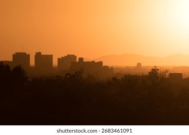 The sun sets behind mountains, casting a fiery orange glow across city skyline. Silhouetted buildings and faint outlines of trees merge with the hazy horizon, creating a dramatic desert sunset scene.