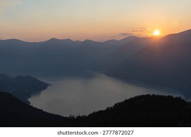 The sun sets behind the mountains, casting soft colors over the lake below. The scene shows hills and water under a sky turning darker as night approaches.