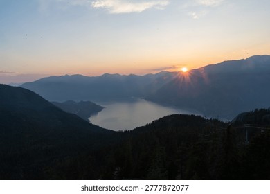 The sun sets behind mountains, casting light over the lake below. The view shows a natural landscape with water and hills during twilight hours.