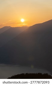 The sun sets behind the mountains, casting long shadows as the sky turns orange. The scene shows the landscape transitioning into evening with a valley visible below.