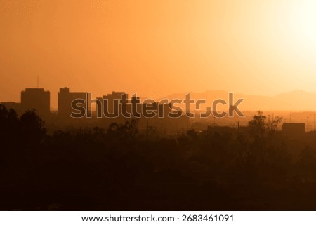 The sun sets behind mountains, casting a fiery orange glow across city skyline. Silhouetted buildings and faint outlines of trees merge with the hazy horizon, creating a dramatic desert sunset scene.