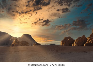 The sun sets behind mountains as clouds gather in the sky. The sandy ground is visible, highlighting the rocky formations. Wadi Rum desert, Jordan, The Valley of the Moon