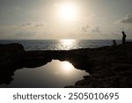 The sun sets behind a rocky coastline. The sunset is reflected in a small lake. On the side, two people look out over the sea.